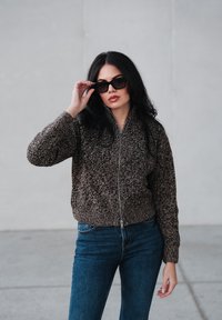 Veste bomber texturée marron et noire avec zip à l'avant, associée à des lunettes de soleil foncées et un jean bleu. Poignets et ourlet lisses et côtelés.