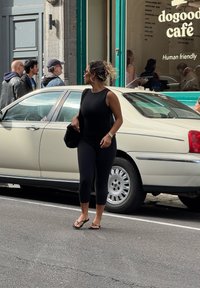 Žena v čiernom športovom oblečení a flip-flopoch stojí na ulici pri béžovom aute, zatiaľ čo traja muži prechádzajú okolo okna kaviarne v pozadí.