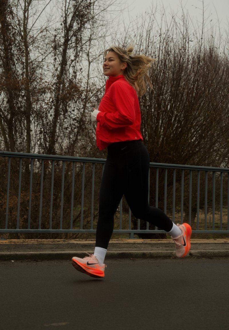 Kvinde løber ude på en grå dag iført en rød jakke, sorte leggings, hvide sokker og pink-orange løbesko.