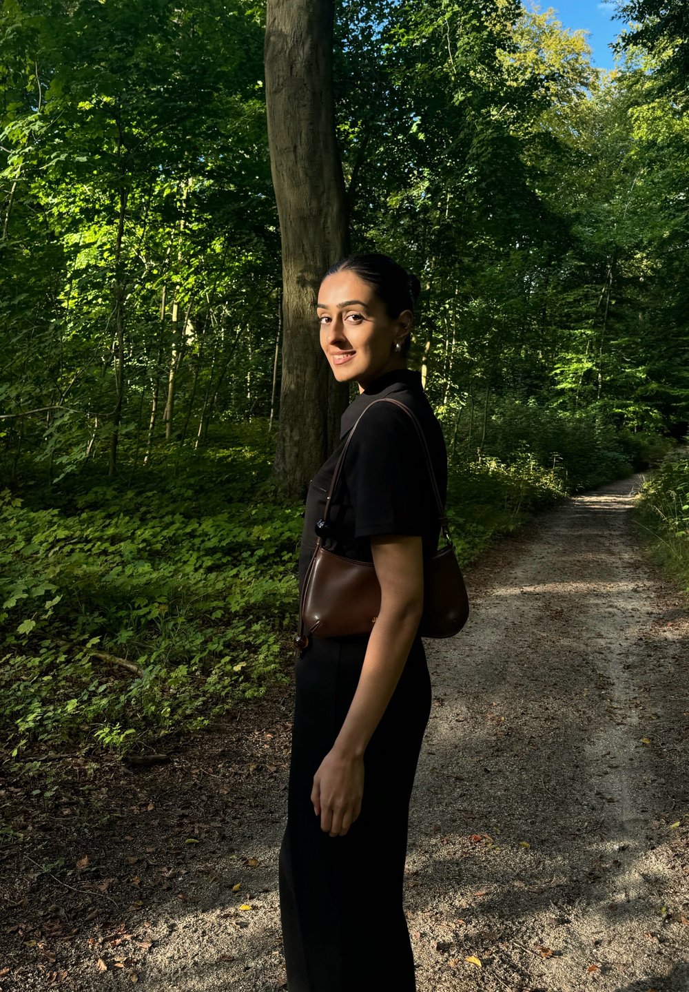 Bruine leren handtas, zwarte kleding, staand op een grindpad omringd door groen en bomen, met zonlicht dat door de bladeren filtert.