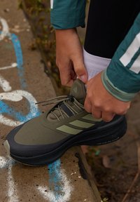 Olivgrüner Sportschuh mit schwarzer Sohle und drei hellgrünen Streifen. Person zieht die Schnürsenkel des Schuhs fest, vor blau-weißem Hintergrund.