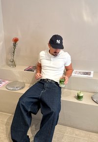 Casual hvid poloshirt, løstsiddende mørkeblå jeans, holder en grøn drik. Minimalistisk baggrund med blomsteraccent og magasiner på overfladen.