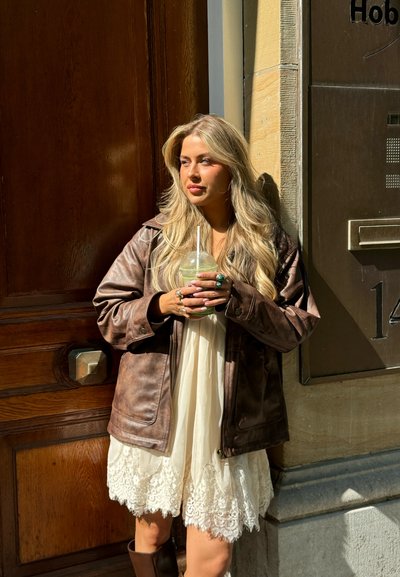 Mujer joven con vestido de encaje blanco y chaqueta de cuero marrón, sosteniendo una bebida con una pajita, de pie junto a una puerta de madera y una pared de piedra bajo la luz del sol.