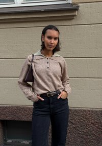 Bruine gebreide trui met een lichte kraag, zwarte high-waisted broek en een kleine bruine schoudertas. Textuurmuur op de achtergrond.