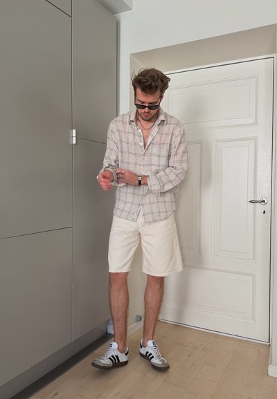 Hombre con gafas de sol, camisa de cuadros, pantalones cortos blancos y zapatillas, remangándose las mangas en el interior junto a una puerta blanca y armarios grises.