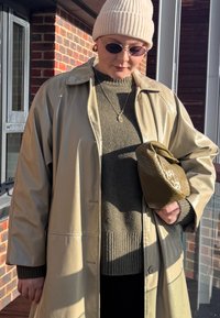 Personne portant un bonnet beige, des lunettes de soleil, un manteau beige brillant et un pull vert, tenant une pochette verte olive texturée à l'extérieur près d'un mur en briques.