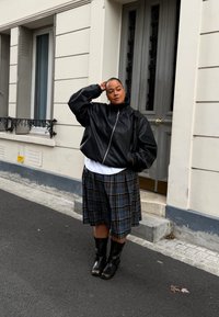 Casaco bomber de couro preto, t-shirt branca, saia plissada em padrão xadrez e botas pretas até ao joelho. De pé contra um edifício com uma porta cinza.