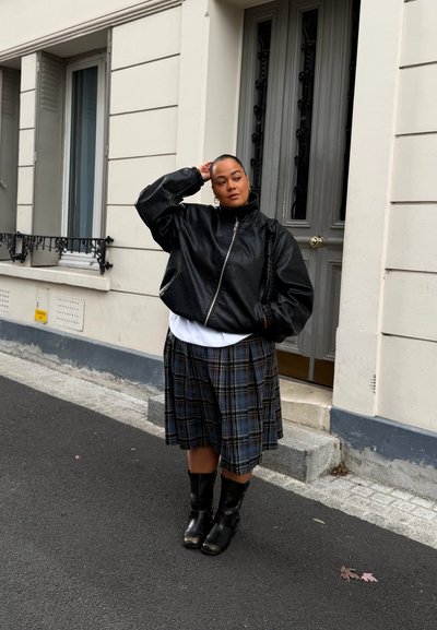 Chaqueta bomber de cuero negro, camiseta blanca, falda plisada de cuadros y botas negras hasta la rodilla. De pie contra un edificio con puerta gris.