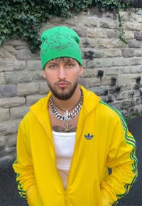Bonnet en tricot vert avec un motif graphique, veste zippée jaune avec des accents bleus, colliers superposés en argent, et un débardeur blanc uni.