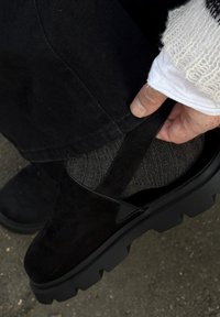 Zwarte suède schoen met een dikke, robuuste zool en elastische zijpaneel, waardoor een gebreide grijze sok eronder zichtbaar is. Een hand past de pasvorm van de schoen aan.