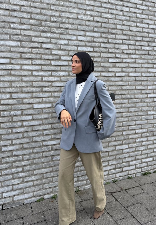 Une femme portant un hijab noir, un blazer gris et un pantalon beige se tient près d’un mur de briques gris et porte un sac à bandoulière à motifs.