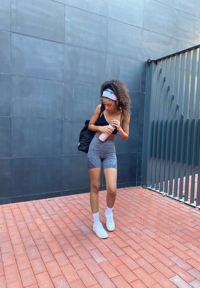 Sujetadora deportiva negra y pantalones cortos grises con estampado, calcetines blancos y zapatillas. Lleva una bolsa negra y una botella de agua rosa, con el pelo rizado y una diadema.