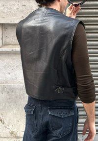 Oseba nosi črno usnjeno jopico čez rjav dolg rokav, temne kavbojke in drži sončna očala blizu svoje glave, z obdano hrbtno stranjo proti kameri.