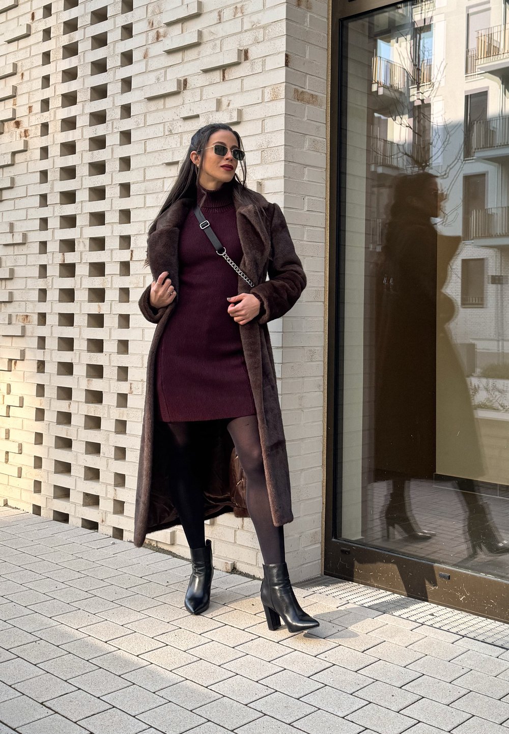 Hnedý umelý kožuch cez burgundskú rebrovú šaty, v kombinácii s čiernymi podpätkovými členkovými topánkami. Model stojí oproti textúrovanej tehlovej stene.