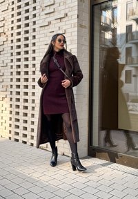 Hnedý umelý kožuch cez burgundskú rebrovú šaty, v kombinácii s čiernymi podpätkovými členkovými topánkami. Model stojí oproti textúrovanej tehlovej stene.