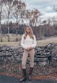 Hvit kabelstrikket genser, beige tettsittende bukser og brune knelange støvler. Står nær en steinmur med trær i landlige omgivelser.