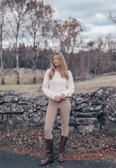 Suéter blanco de punto con cable, pantalones ajustados beige y botas marrones hasta la rodilla. De pie cerca de un muro de piedra con árboles en un entorno rural.