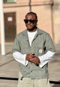 Mężczyzna w okularach przeciwsłonecznych, w zielonej, warstwowej koszuli z krótkim rękawem na białej koszulce z długim rękawem, z kilkoma pierścieniami i naszyjnikami, stojący na zewnątrz na słońcu.