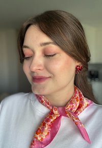 Orecchini a forma di fragola rossa con pois bianchi, abbinati a una sciarpa con motivo floreale rosa e a un morbido pullover bianco.