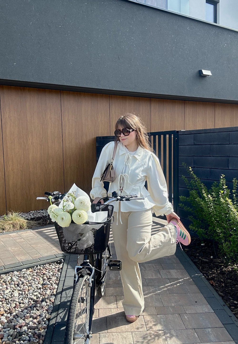 Vit blus med puffärmar, beige byxor och rosa skor. Svart cykel med en nätkorg fylld med vita blommor och grönska.