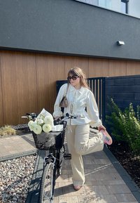 Vit blus med puffärmar, beige byxor och rosa skor. Svart cykel med en nätkorg fylld med vita blommor och grönska.