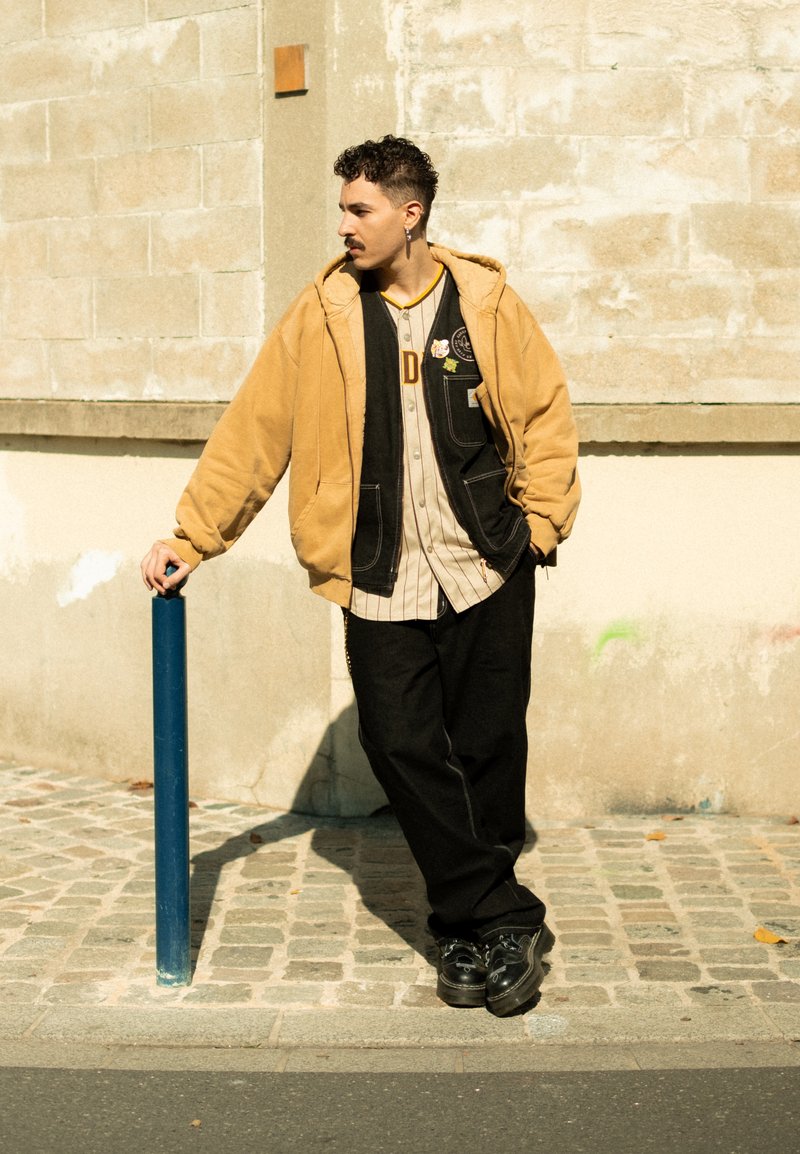 Eine Person lehnt an einem blauen Pfahl und trägt einen beigen Hoodie über einem schwarz-weißen gestreiften Hemd, schwarze Hosen und schwarze Schuhe.