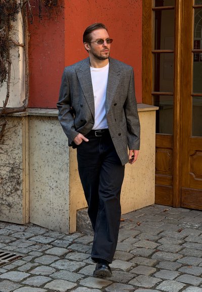 Hombre con blazer de cuadros gris, camisa blanca, pantalones negros y gafas de sol caminando sobre adoquines cerca de una pared naranja y una puerta de madera.