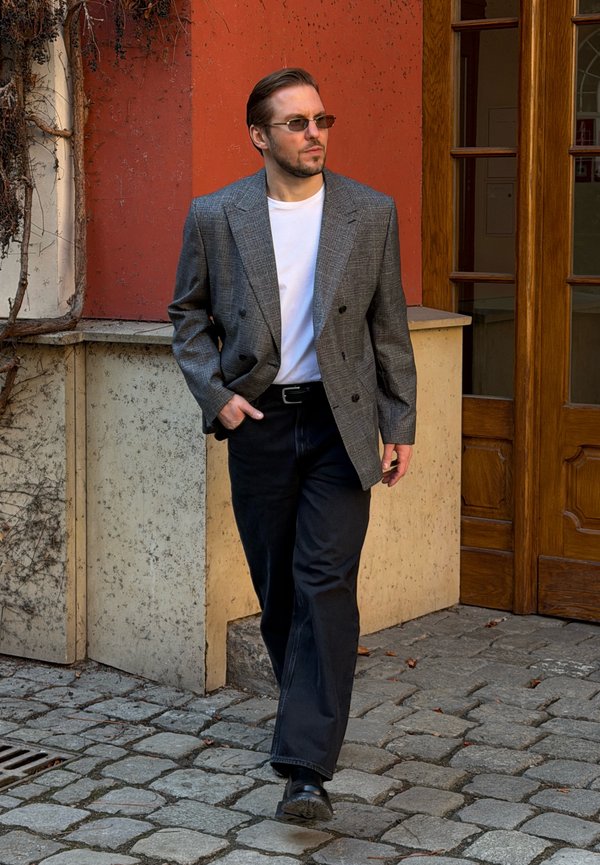 Άνδρας που φοράει γκρι καρό σακάκι, λευκό πουκάμισο, μαύρο παντελόνι και γυαλιά ηλίου, περπατώντας σε πλακόστρωτο κοντά σε πορτοκαλί τοίχο και ξύλινη πόρτα.