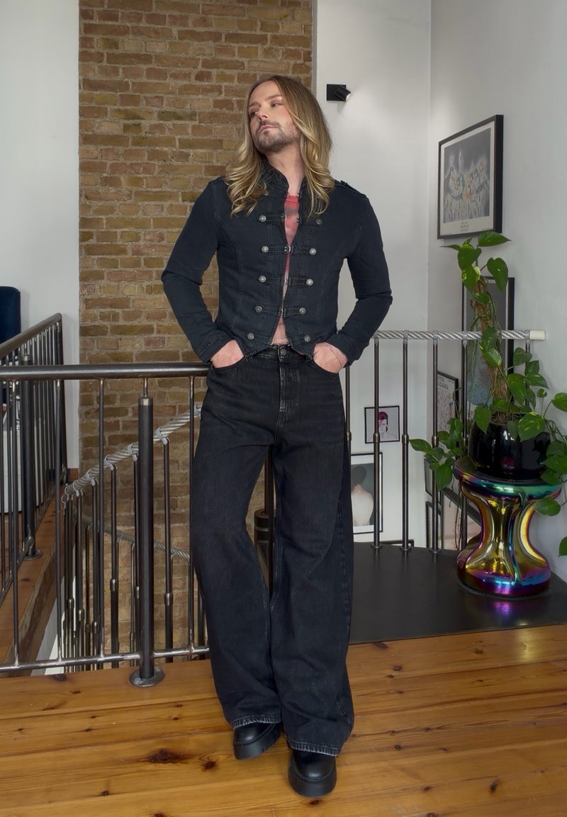 Personne aux longs cheveux blonds debout à l'intérieur portant une veste noire courte, un jean noir large et des chaussures à plateforme noires, les mains dans les poches.