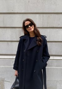 Marineblå dobbeltspent kåpe med store knapper, sidelommer og belte. Glatt ullstoff med skreddersydd design og skulderdetaljer.