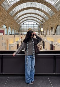 Paksu harmaa neulepusero, jossa leveät hihat, siniset farkut ja kuvioitu huivi. Ruskea käsilaukku. Taustalla modernin museon sisätila.