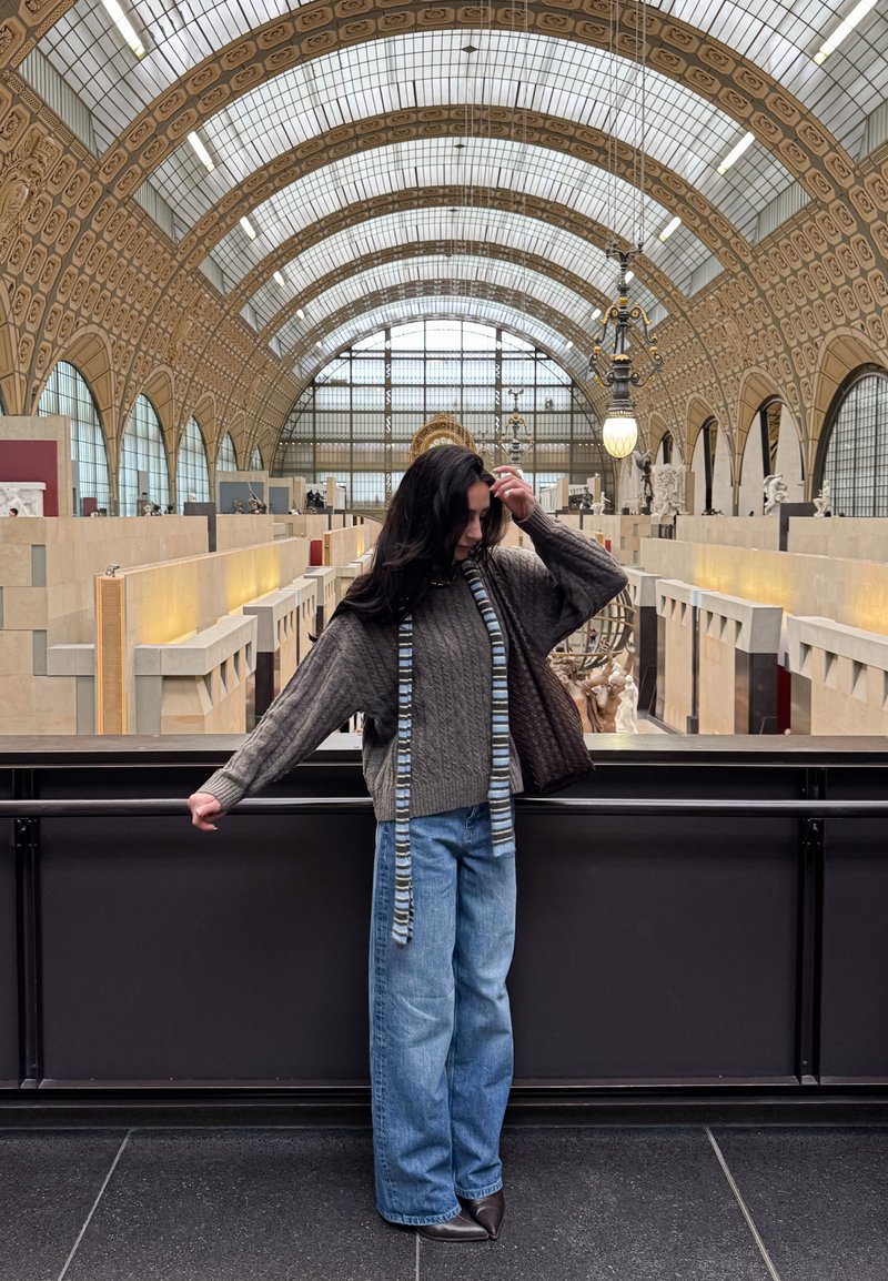 Dicker grauer Strickpullover mit weiten Ärmeln, blaue Jeans und gemusterter Schal. Braune Handtasche. Modernes Museuminterieur im Hintergrund.