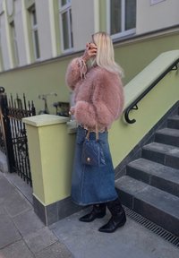 Femeie cu păr lung și blond, purtând o jachetă roz din blană, fustă din denim, cizme negre și ținând o geantă albastră matlasată, aproape de scări.