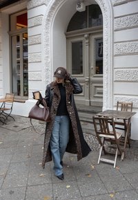 Casaco preto longo com forro de estampado de leopardos, combinado com um suéter escuro e jeans azuis. Bolsa castanha e chávena de café na mão, de pé no passeio.