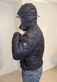 Άτομο με μπλε τζιν ρυθμίζει τη κουκούλα ενός μαύρου φουσκωτού μπουφάν σε εσωτερικό χώρο, στραμμένο προς τα αριστερά, με λευκό καπέλο κάτω από την κουκούλα.