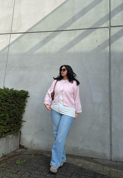 Mujer con el pelo largo y oscuro que lleva gafas de sol, un cárdigan rosa, camisa blanca, vaqueros azul claro y zapatos beige, de pie junto a una pared de hormigón gris.