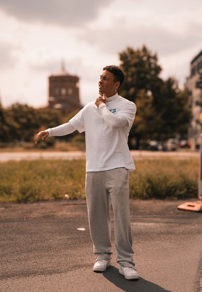 Camiseta blanca de manga larga, pantalones de chándal grises y zapatillas blancas. De pie sobre asfalto con hierba verde al fondo y un edificio detrás.