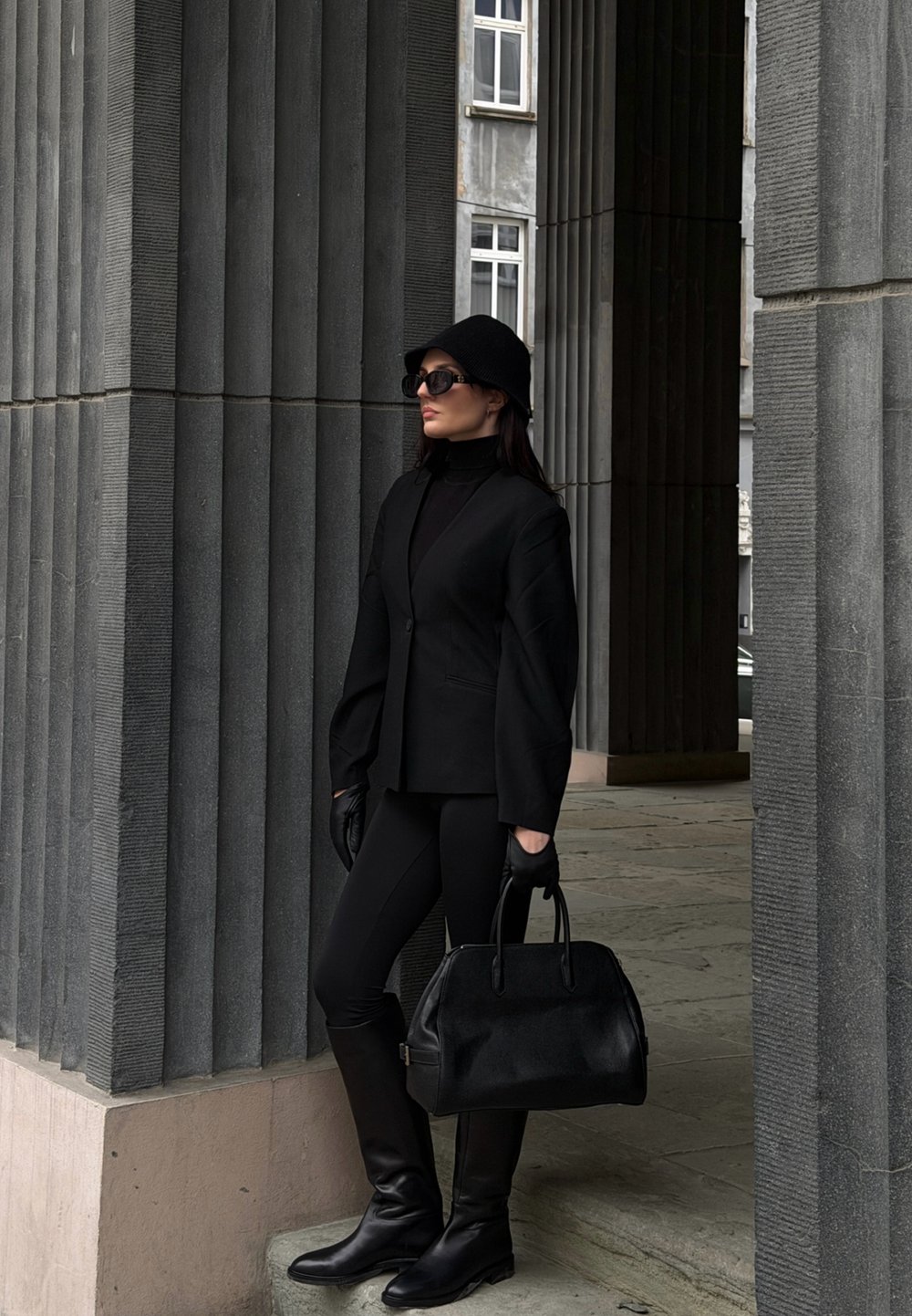 Sort skræddersyet blazer med brede ærmer, sort rullekrave, læderhandsker, tætsiddende sorte leggings, knælange sorte støvler og en struktureret håndtaske.