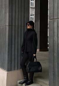 Sort skræddersyet blazer med brede ærmer, sort rullekrave, læderhandsker, tætsiddende sorte leggings, knælange sorte støvler og en struktureret håndtaske.