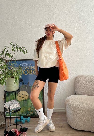 Camiseta blanca de gran tamaño, pantalones cortos negros, bolsa de hombro naranja, calcetines blancos con el logo de Nike y zapatillas blancas. Entorno interior con plantas y arte.