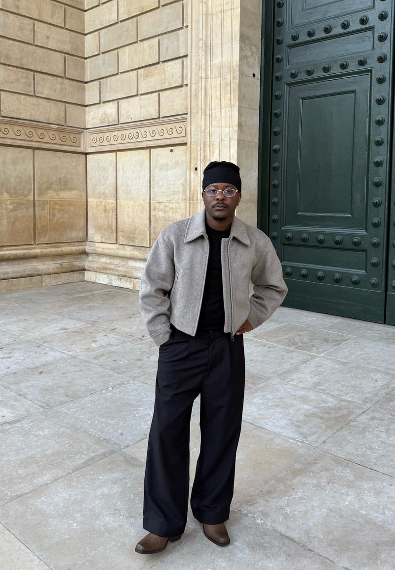Veste gris en mélange de laine avec fermeture éclair, t-shirt noir, pantalons larges noirs et bottines marron à enfiler. Porter des lunettes transparentes et un bonnet noir.