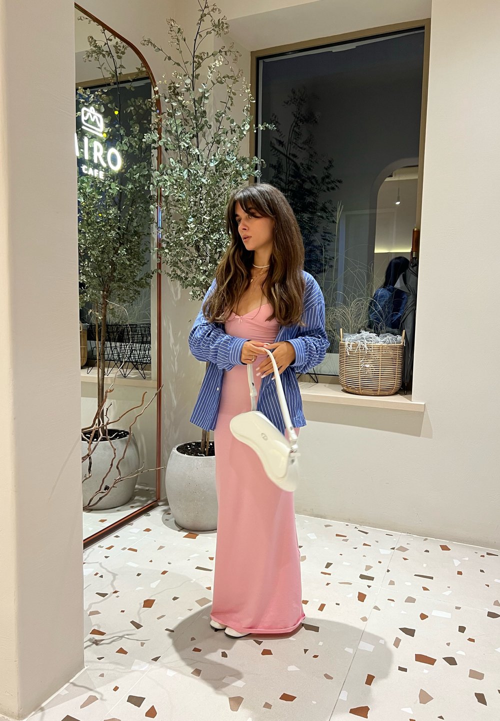 Modèle dans une robe rose ajustée avec des bretelles spaghetti, superposée avec une chemise à rayures bleues. Tient un sac à main blanc, debout sur un sol à motifs.