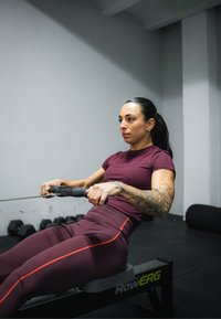 Rowing machine with a user. The machine is black, featuring the "RowERG" logo. The user is wearing a burgundy top and sports leggings with orange accents.