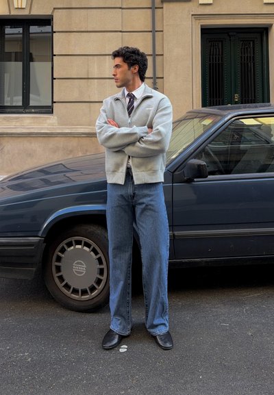 Joven con los brazos cruzados está de pie en la calle junto a un coche Volvo vintage azul oscuro frente a un edificio beige con ventanas y puerta.