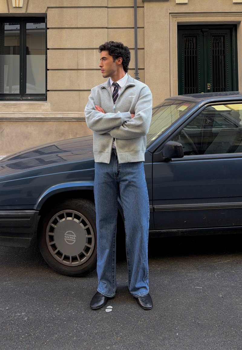 Jeune homme les bras croisés, debout dans la rue à côté d'une voiture vintage Volvo bleu foncé devant un bâtiment beige avec fenêtres et porte.