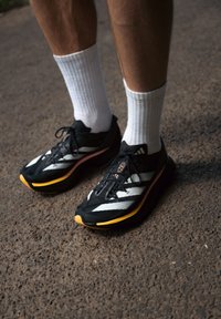 Des chaussures de course noires légères avec des rayures blanches, une semelle jaune et une texture en maille, associées à des chaussettes blanches au niveau de la cheville sur une surface en gravier.