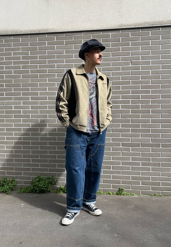 Man met zwarte hoed, beige jas met zwarte strepen, grafisch T-shirt, wijde jeans en zwarte sneakers staat bij een bakstenen muur in het zonlicht.