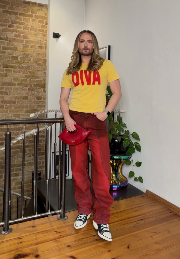 Un hombre de pelo largo está de pie junto a una barandilla. Lleva una camiseta amarilla con la palabra «DIVA», pantalones rojos y zapatillas deportivas, y sostiene un bolso de mano rojo.
