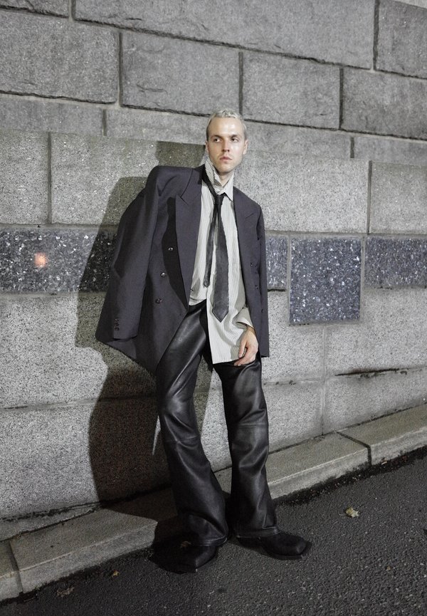 Jonge man met kort blond haar leunt tegen een stenen muur, draagt 's nachts een oversized zwarte blazer, gestreept hemd, zwarte das en brede leren broek.