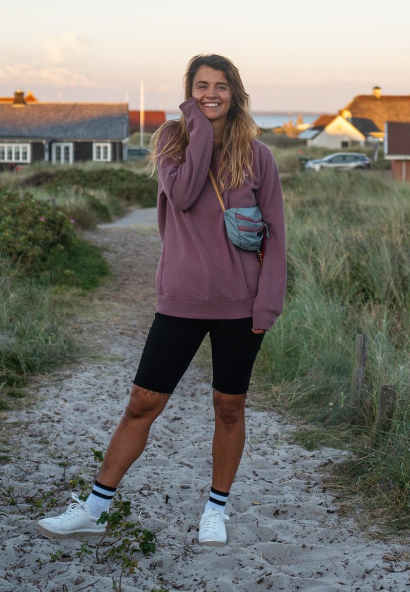 Felpa viola, pantaloni corti neri, sneakers bianche con calzini a righe nere e blu, e una piccola borsa a tracolla grigia. Sentiero di sabbia, erba sullo sfondo.